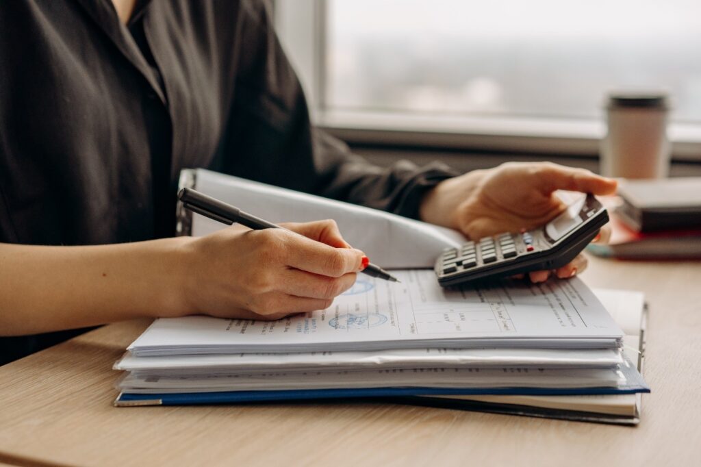 A person holding a calculator in one hand and a pen in the other to record something in a file.